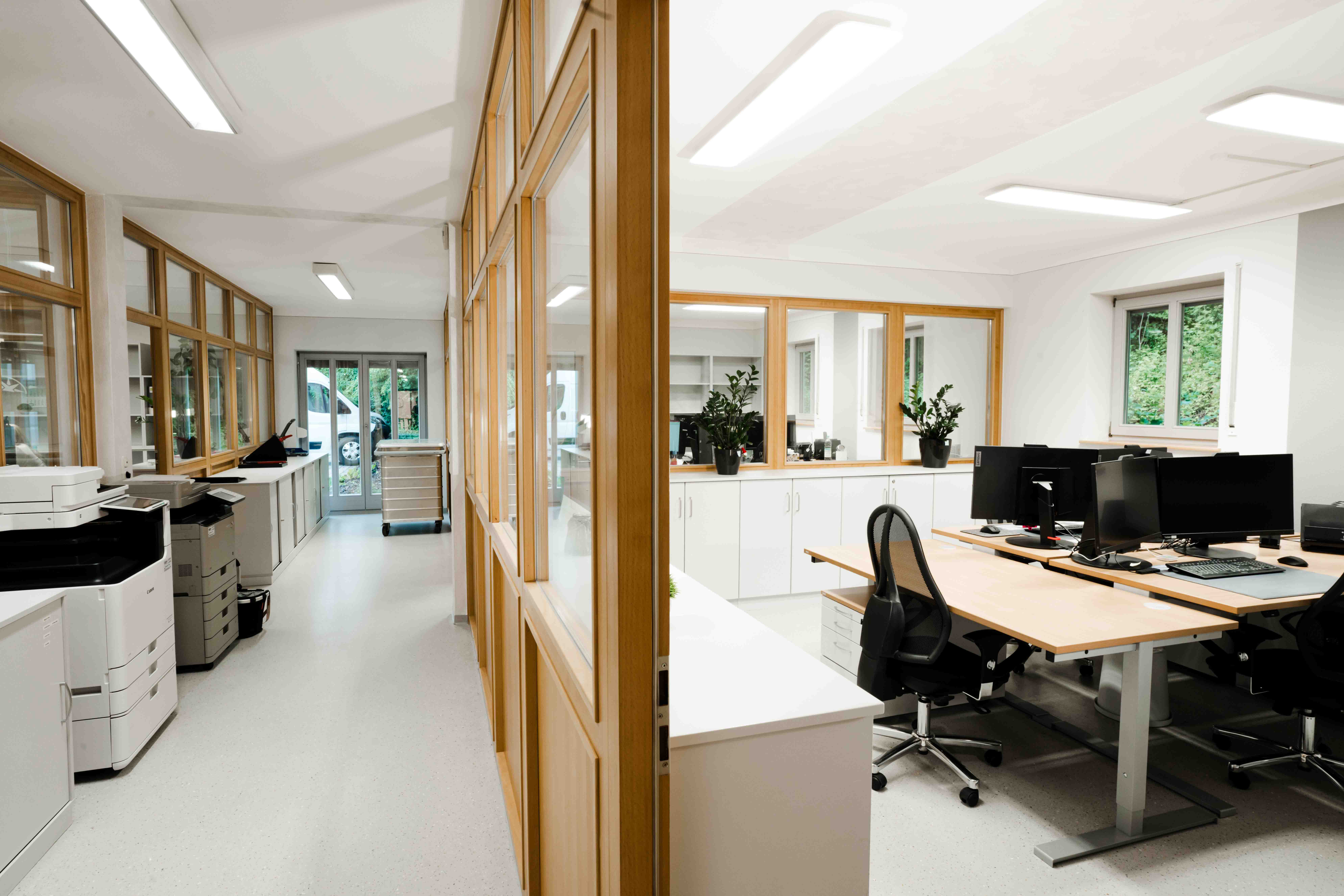 Büroraum mit Holz- und Glastrennwänden, Durchgang, Drucker, Sideboards, rechts Büro mit Schreibtischen, Monitoren und Stühlen.
